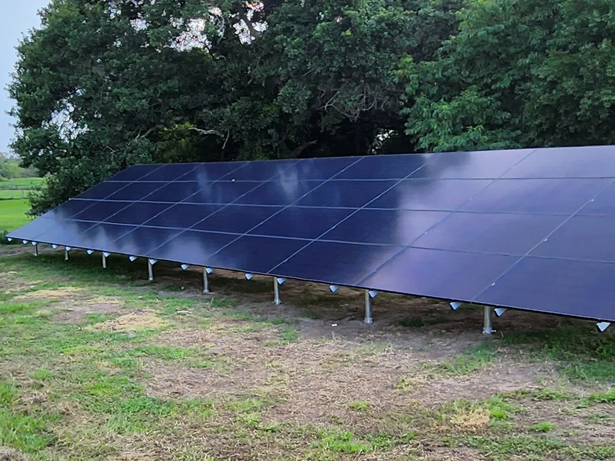 Residential ground-mounted solar panel array installed on a reinforced steel frame with a tree line in the background.