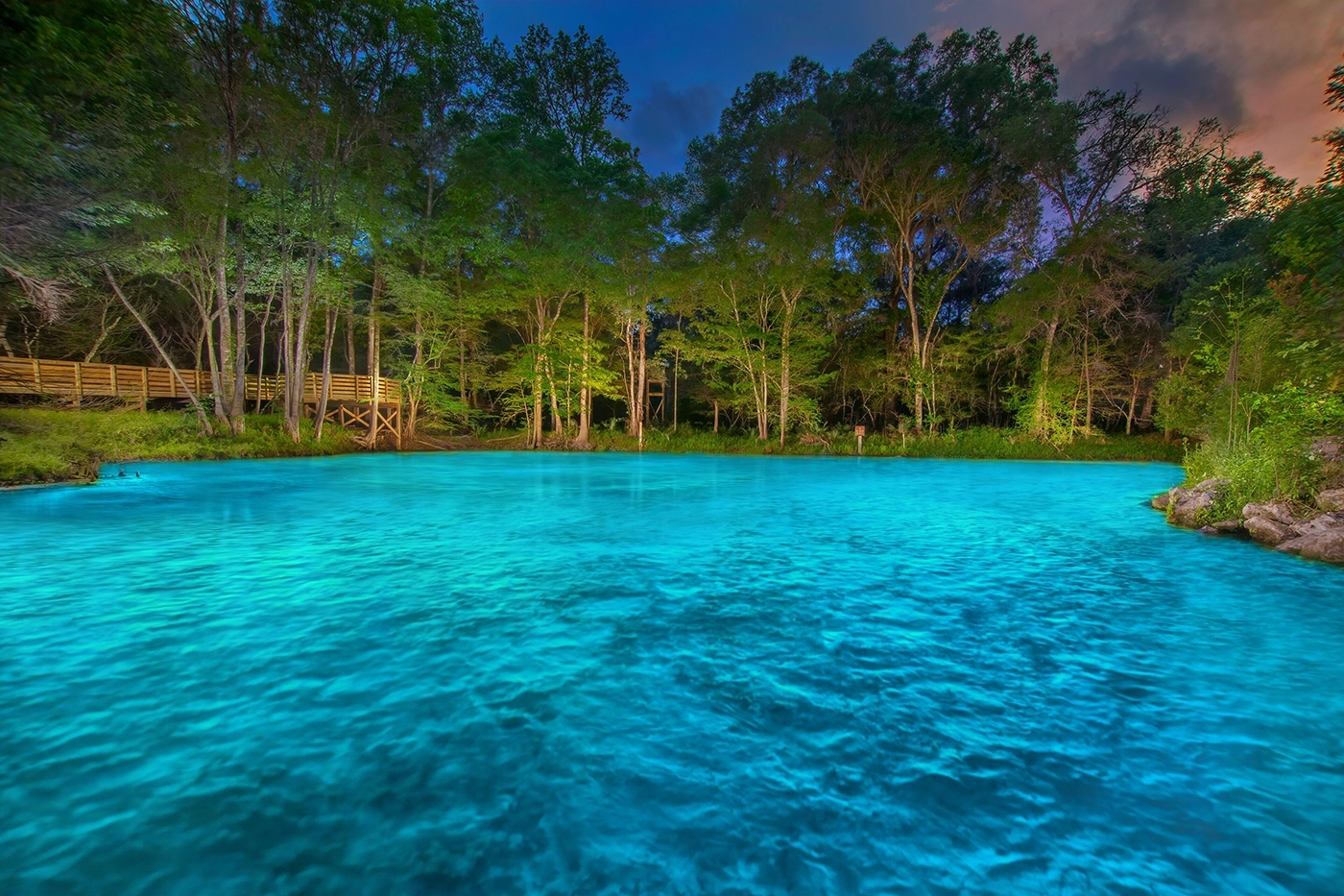 Turquoise lake edged by a dense forest with a wooden boardwalk along the left shore at dusk.