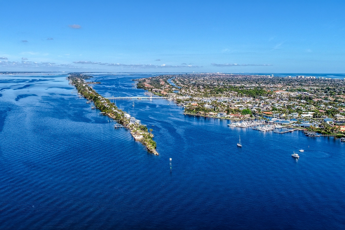 Aerial view of a coastal town with a long narrow island lined with houses stretching into blue water and boats docked along the shoreline.