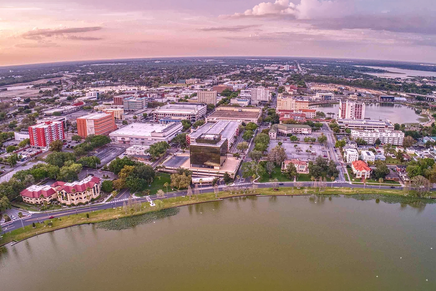 Aerial view of a riverfront city at sunset, with a mix of low-rise buildings, trees, and a curved road along the water.
